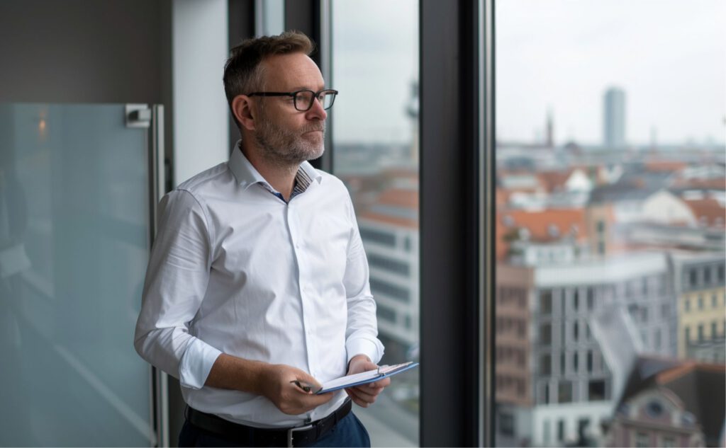 Nachdenklicher GmbH-Geschäftsführer Ende 30 steht am hohen Fenster eines modernen Büros in Berlin Mitte mit Blick über die Stadt, hält einen gefalteten Vertrag und wägt eine Entscheidung ab