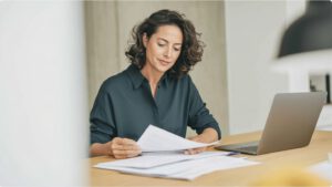 Frau sitzt am Schreibtisch, haelt ein Schreiben in der Hand, Laptop daneben